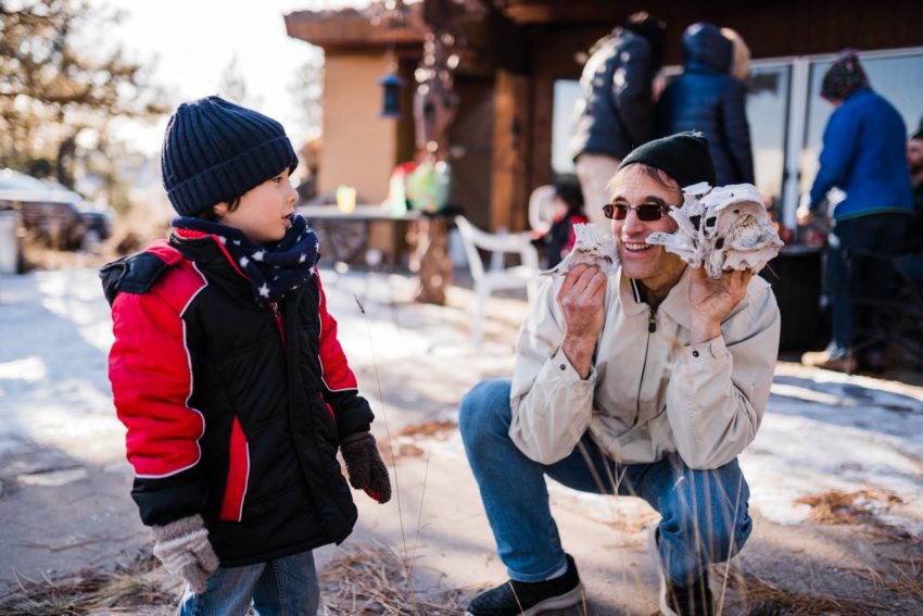Child in blue beanie and red-black jacket on the left; woman in a beige coat crouches with a small light-colored dog in a snowy outdoor scene.