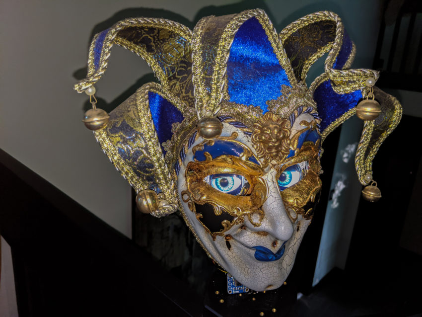 Ornate blue-and-gold jester mask with gold filigree, blue fabric panels, bells at the tips, and a white face with blue iris detail.