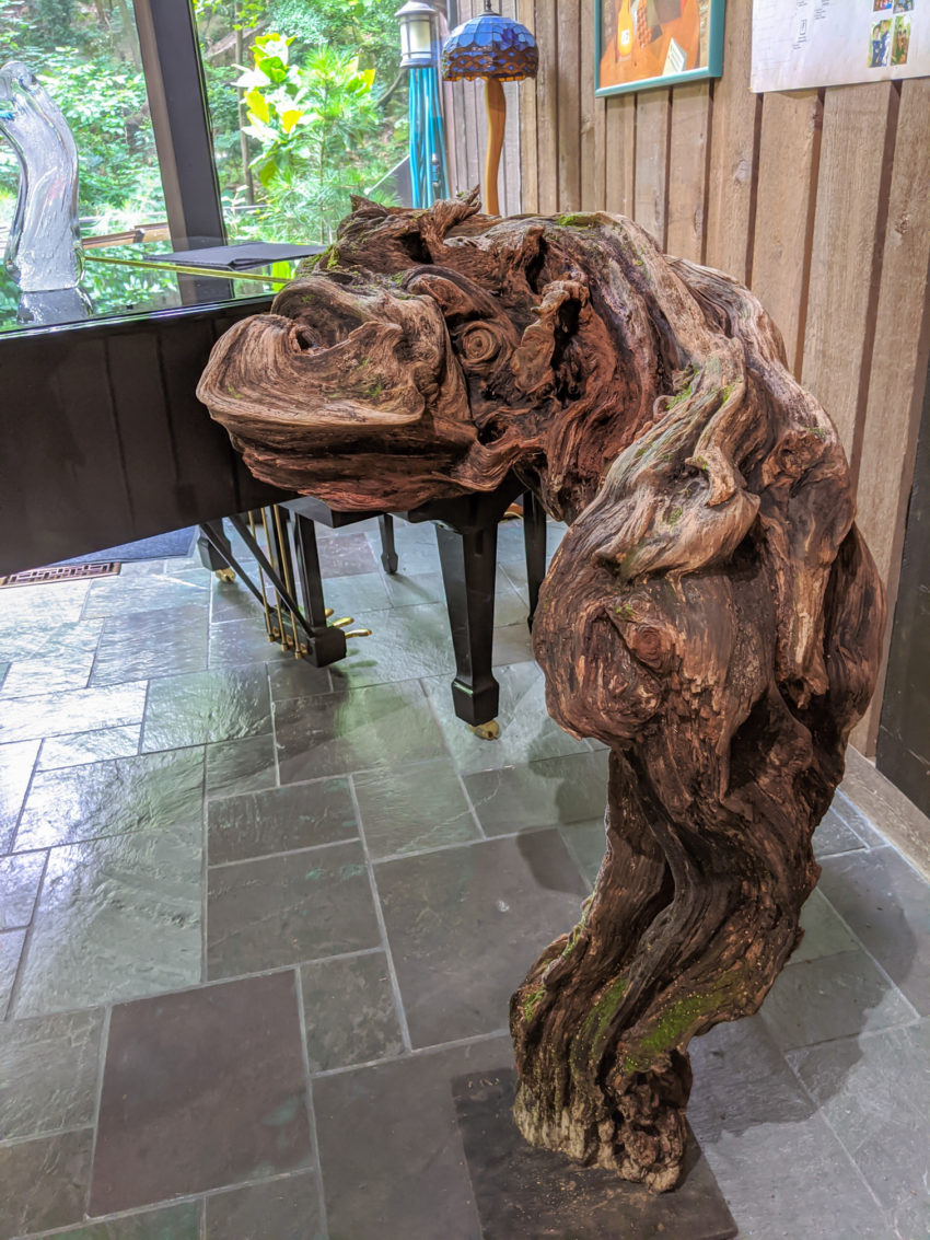 Brown carved wooden four-legged creature with a long snout, indoors on a tiled floor near wood-paneled walls and framed art.