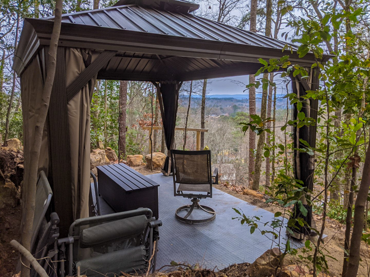 Dark gazebo with curtains on a rocky hilltop deck; two chairs and a swivel chair overlook a forested valley.