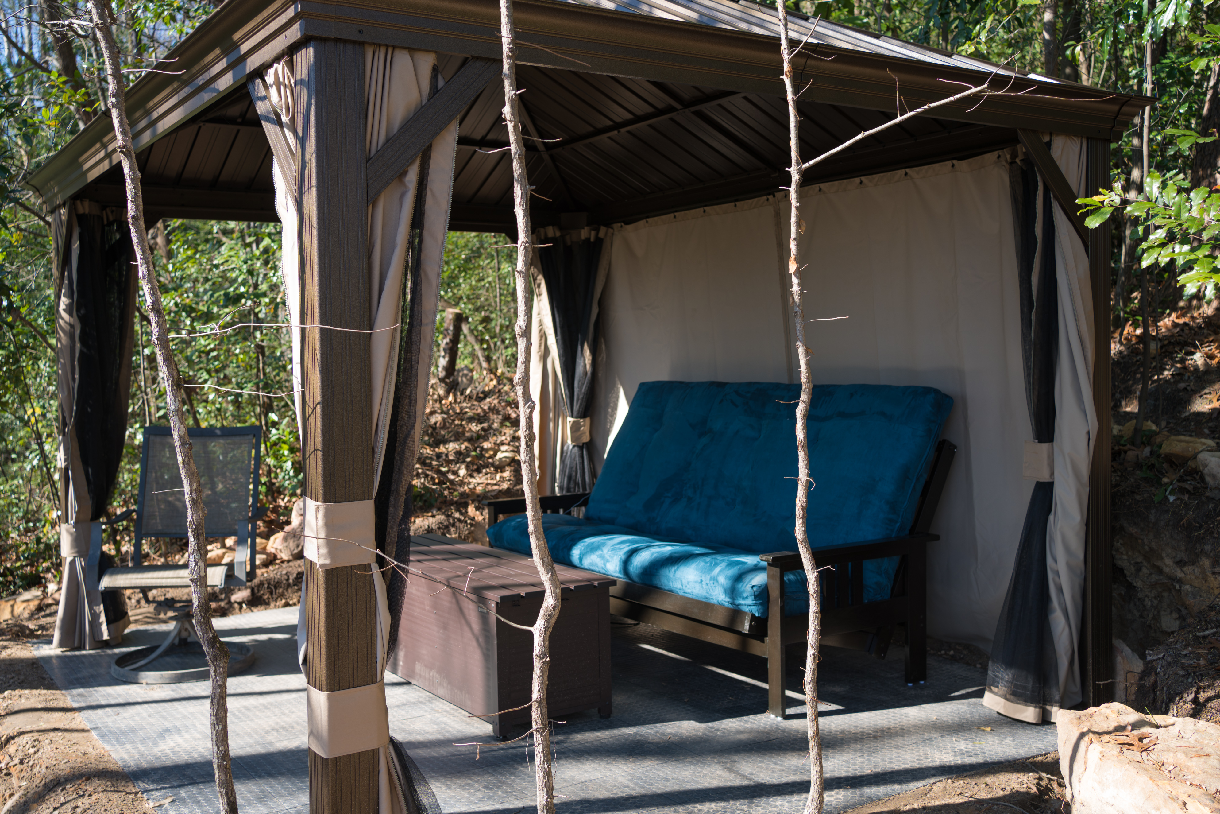 Outdoor gazebo with beige curtains and brown frame, blue cushioned sofa, dark brown coffee table on a metal deck, surrounded by trees.