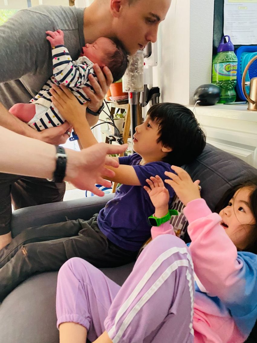 Man leaning over a gray sofa kissing a striped baby; two kids in purple and pink watch in a bright living room.