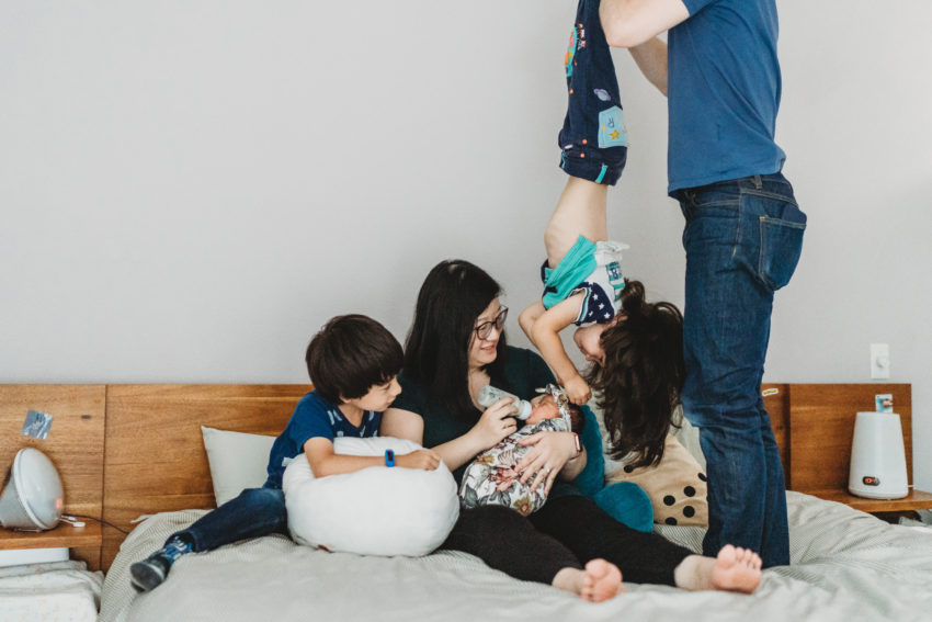 Mother and two kids sit on white bedding; a person in jeans stands at the edge. Wooden headboard and small lamps on nightstands.