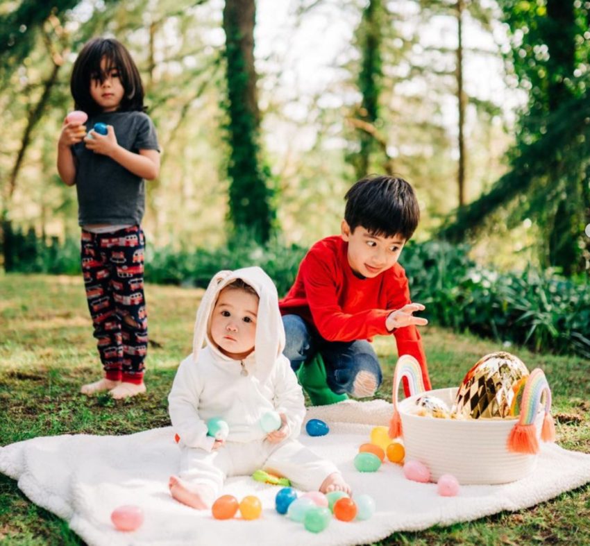 Children on a white blanket on the grass, with colorful toys; a red-shirted child sits beside a white-hooded child, another stands nearby.