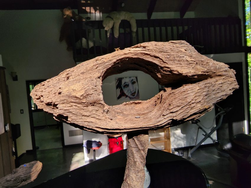 Rough brown ring sculpture on a pedestal, hollow center frames a small black-and-white portrait; gallery interior in background.