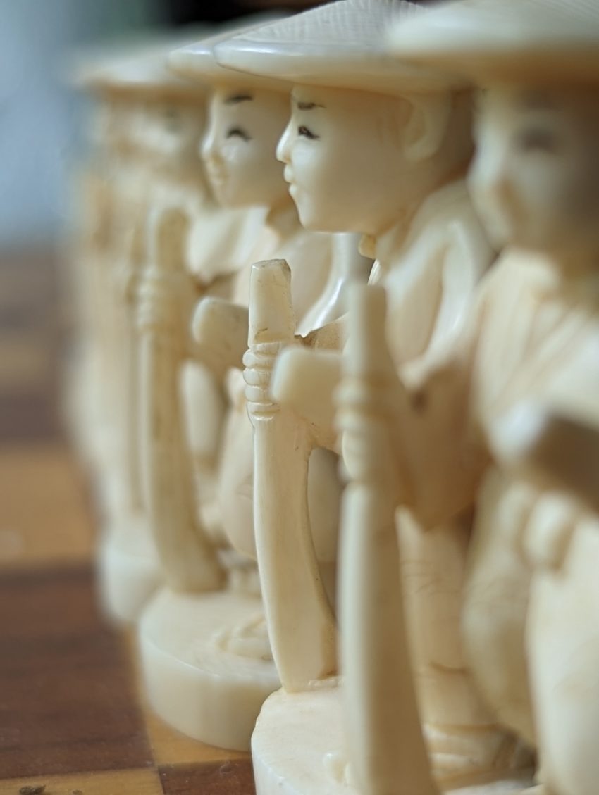 Row of cream-colored wooden figurines with hats lined up on a wooden chessboard, shallow depth of field.