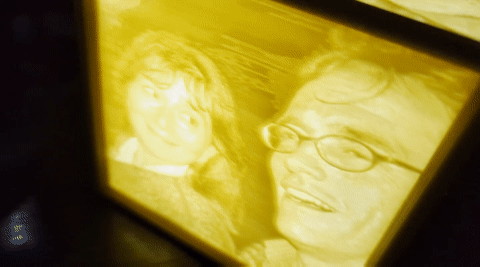 Rectangular frame lit with yellow light, showing two portraits: left a young person, right an older man with glasses.