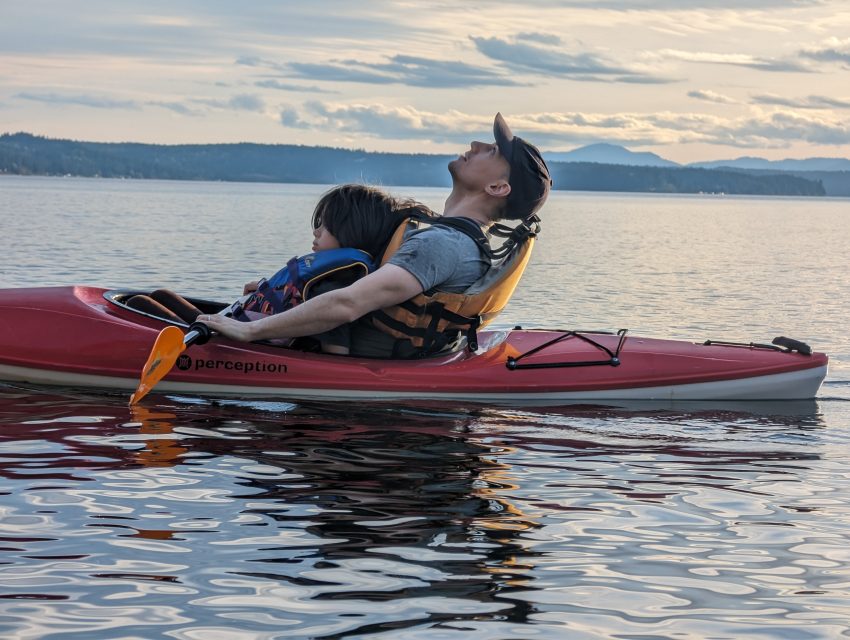 Red tandem kayak on calm water with two people in life jackets; one leans back wearing a cap, cloudy horizon and distant hills.
