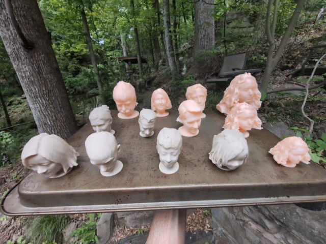 Array of small clay busts and heads in peach and gray on a wooden table outdoors among trees.
