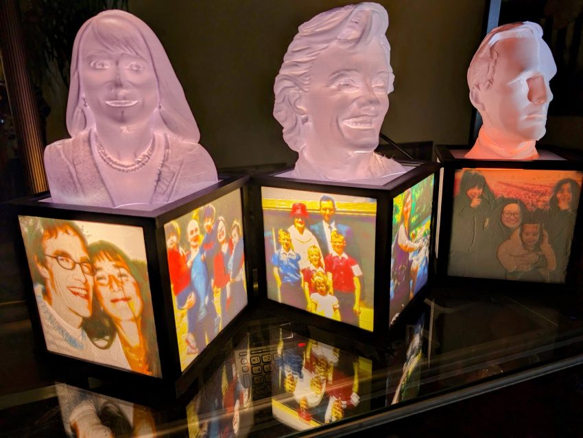 Three pink busts atop black-framed photo lanterns with family pictures on their sides, lined up on a glass shelf with reflections.