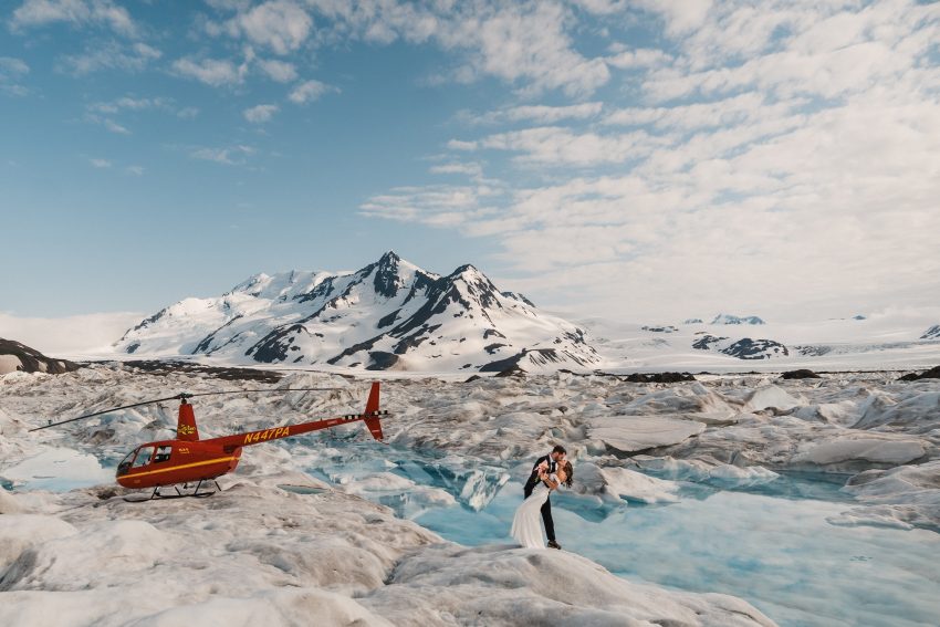Bride and groom on glacier beside red helicopter and turquoise meltwater, snow-covered mountains under blue sky