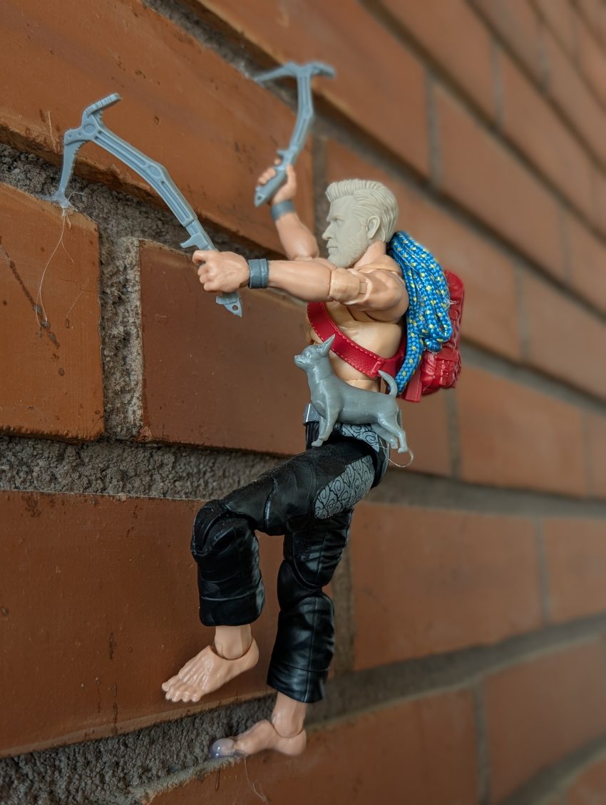 Toy action figure climbs a brown-orange brick wall using light-blue cords; red top, black pants, bare feet, blue backpack.
