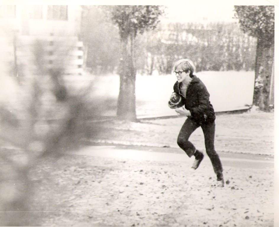 Black-and-white scene: a person with glasses runs along a leaf-strewn street, cradling a small bundled child.