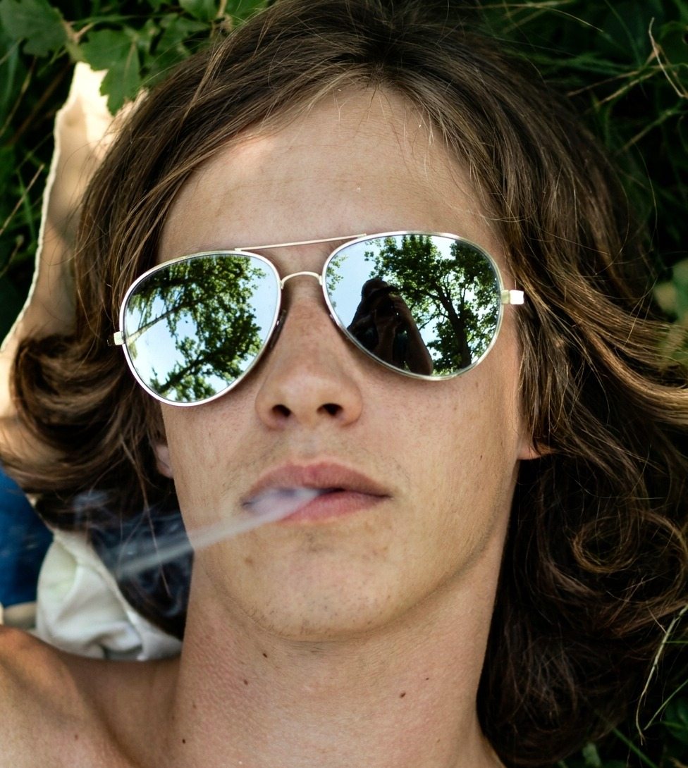 Person with long hair wearing aviator sunglasses lies in grass, smoking; mirrored lenses reflect trees and a photographer.